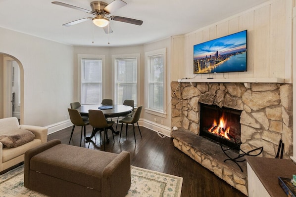 A living room with a stone fireplace, a television mounted above it, and a dining area with a round table and chairs.