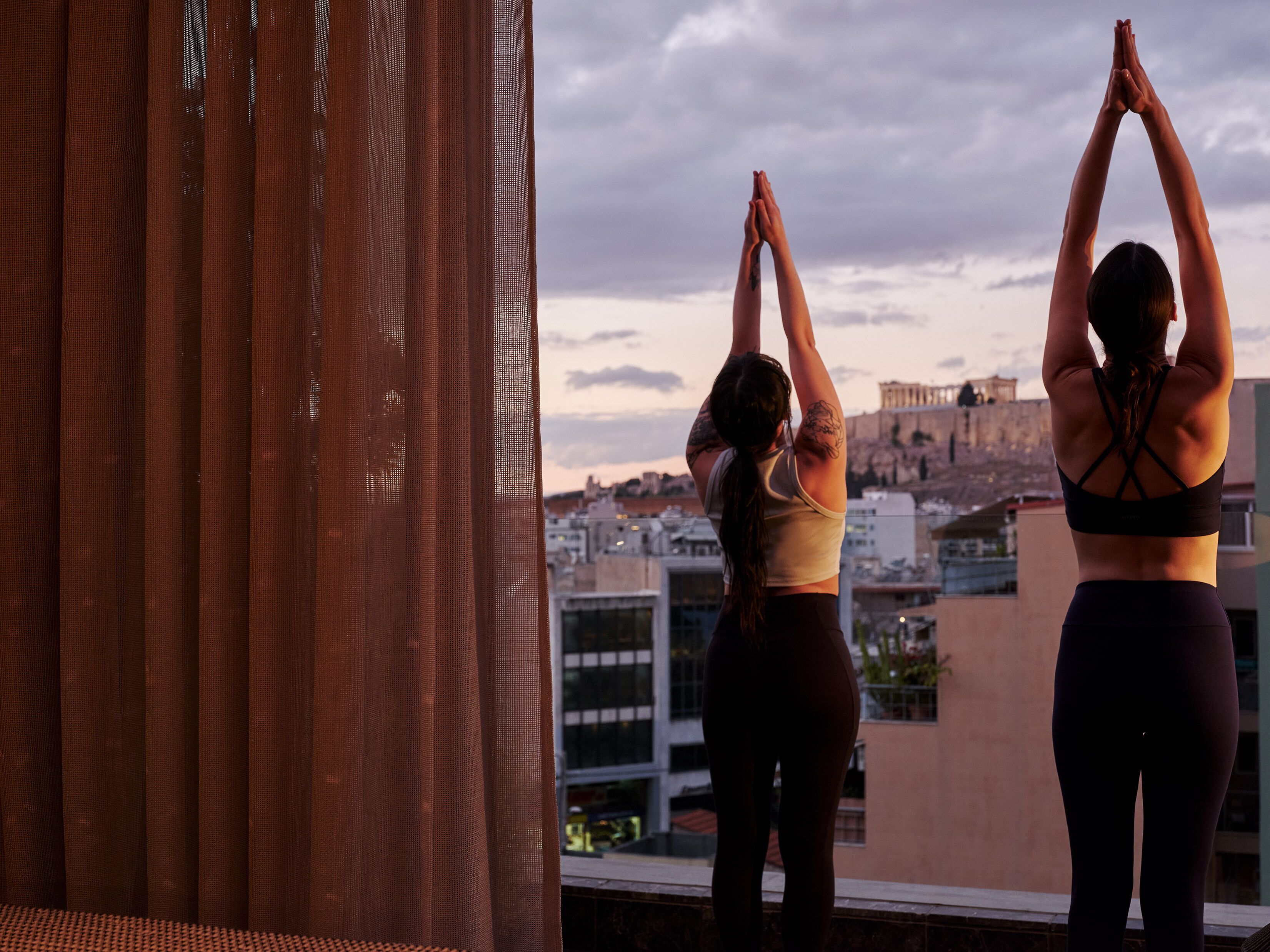 Two individuals performing yoga on a balcony with a cityscape and ancient ruins in the background.