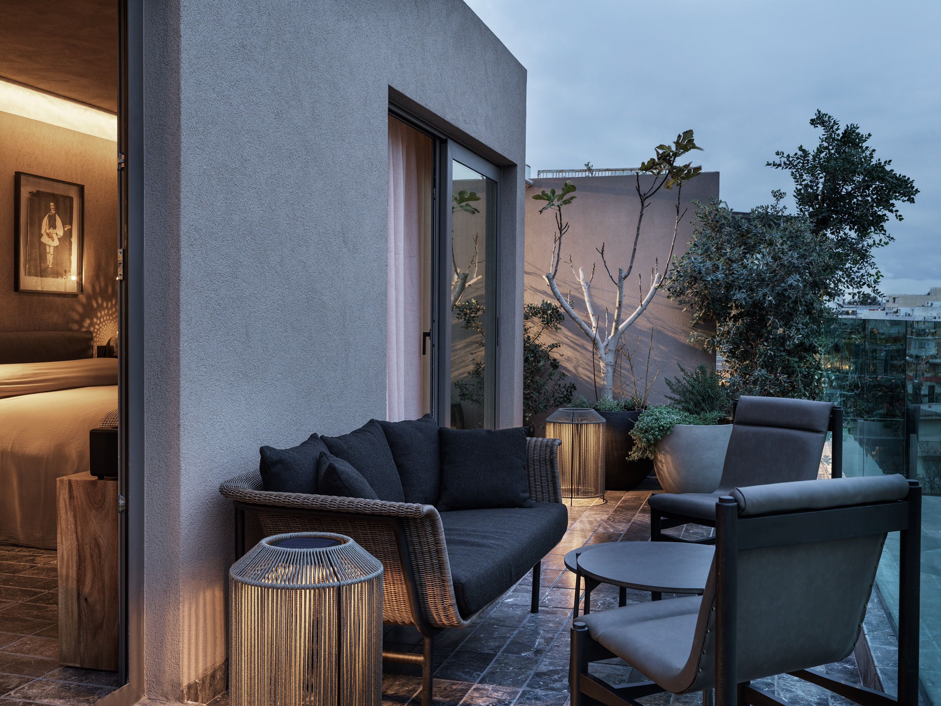 A modern outdoor seating area with a sofa, chairs, and a small table, featuring a view of the cityscape and a tree.