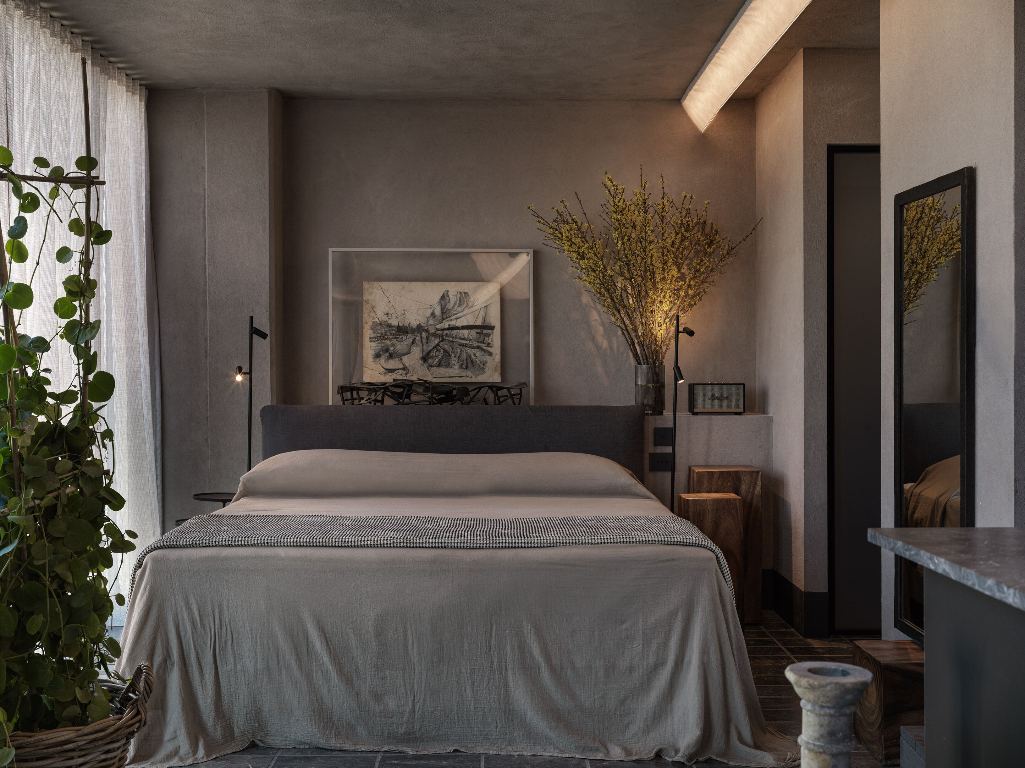 A modern bedroom with a large bed, a bedside table, a floor lamp, a framed artwork, and a vase with branches.