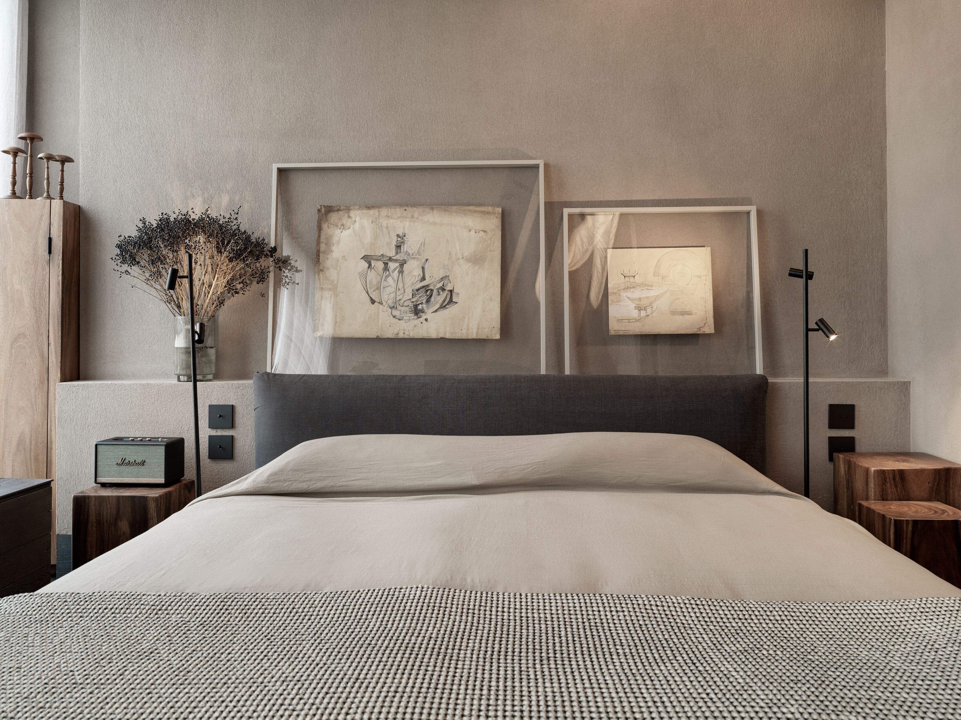 A modern bedroom with a large bed, two framed artworks on the wall, a vase with dried flowers, a Marshall speaker, and a tall floor lamp.