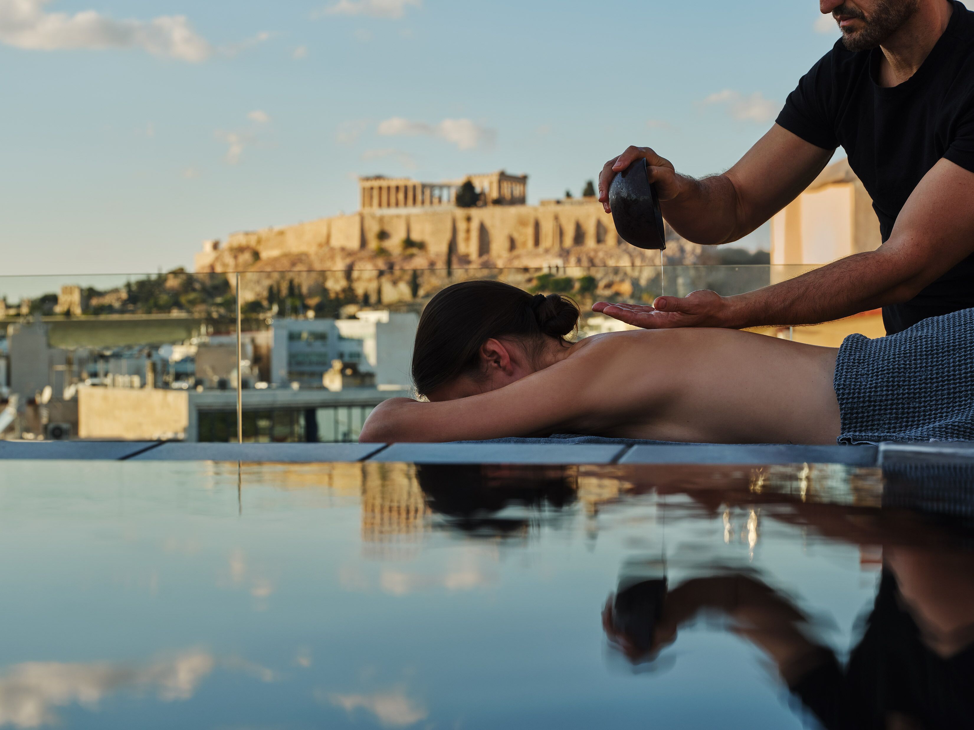 A person receiving a back massage on an outdoor pool with a view of ancient architecture in the background.