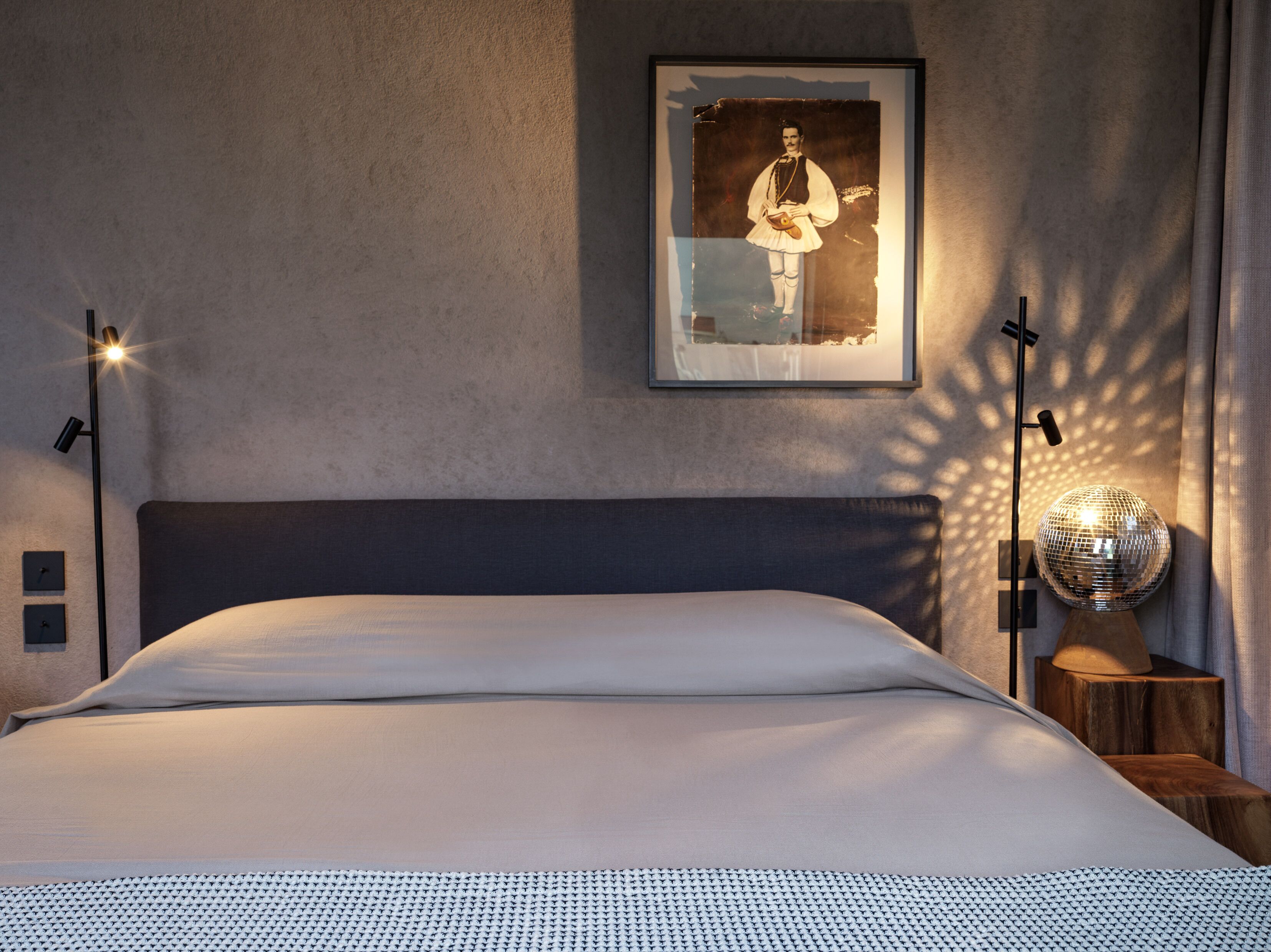 A neatly made bed with a patterned bedspread, a framed picture on the wall, and two floor lamps.