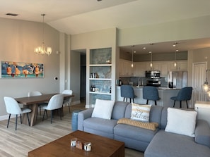 A modern living room with a grey sofa, a wooden coffee table, and a dining area with a wooden table and chairs. A kitchen with white cabinets and stainless steel appliances is visible in the background. - Serenata - Beautiful Luxury, Top Floor Corner, Water View! (Sarasota)