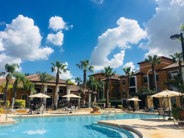 A swimming pool with lounge chairs and umbrellas, surrounded by palm trees and buildings with tiled roofs. - Serenata - Beautiful Luxury, Top Floor Corner, Water View! (Sarasota)