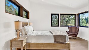 Ein Schlafzimmer mit einem hölzernen Bett, einem Nachttisch, einem Sessel und großen Fenstern mit Blick auf Bäume.
