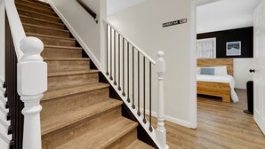 A staircase with wooden steps and white balusters, leading to a bedroom with a bed and a nightstand. - House on The Rocks with EV Charger (Chittenden)