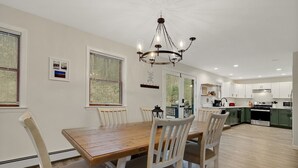 A dining area with a wooden table and white chairs, a chandelier, and a kitchen in the background. - House on The Rocks with EV Charger (Chittenden)