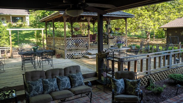 Un patio techado al aire libre con cubierta de madera, zona de estar con sofás y sillones acolchados, y un pabellón con techo de celosía.