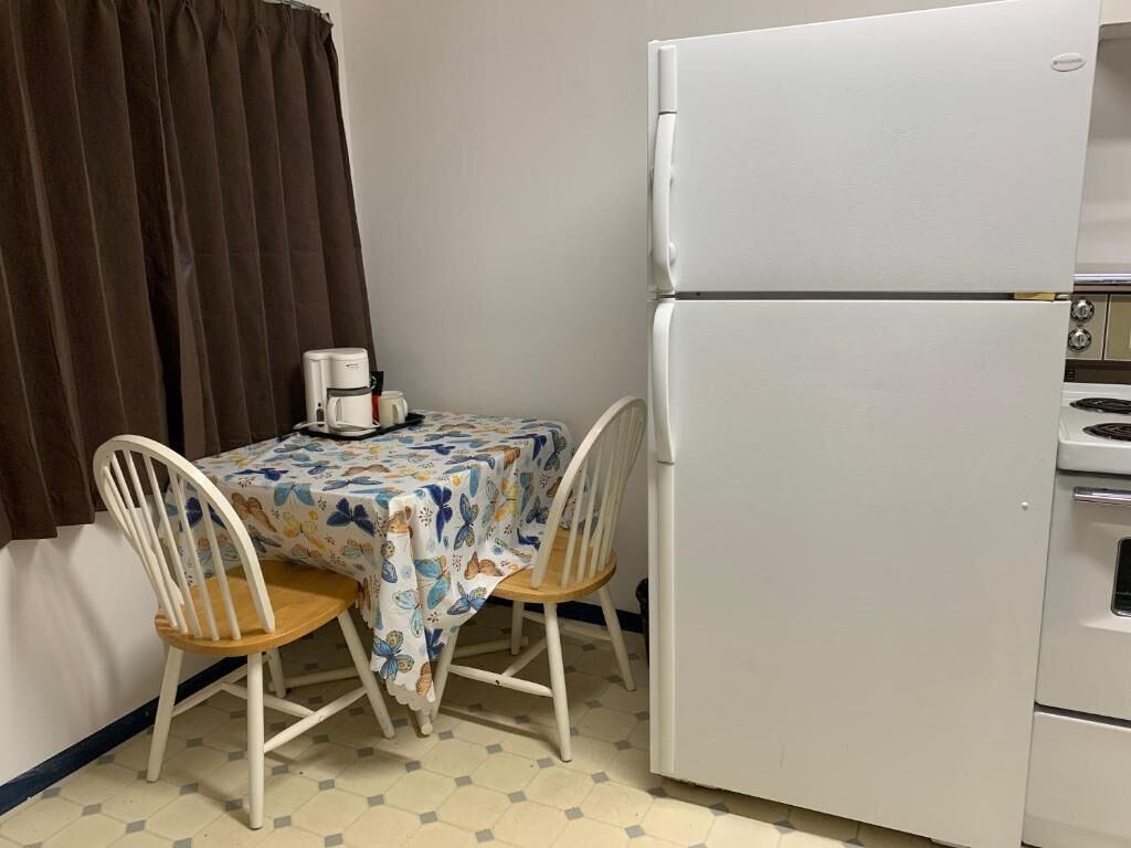 Une cuisine compacte avec un réfrigérateur blanc, une petite table à manger recouverte d’une nappe à motifs floraux, deux chaises en bois et une machine à café.