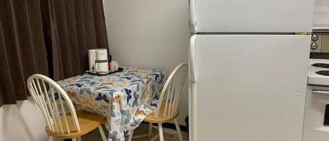 Une cuisine compacte avec un réfrigérateur blanc, une petite table à manger recouverte d’une nappe à motifs floraux, deux chaises en bois et une machine à café.
