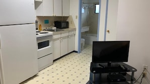 A compact kitchen with white cabinets, a stove, and a microwave. There's a black TV stand with a TV and remote. A shower is visible through a glass door.