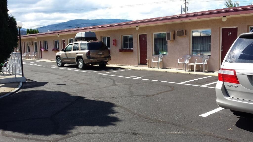 Un motel de una sola planta, habitación número 23, con aparcamiento, dos coches, zona de estar al aire libre y vistas a la montaña al fondo.