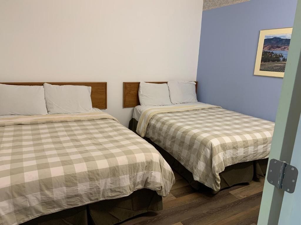 Une chambre avec deux lits, chacun recouvert d’une courtepointe à carreaux et agrémenté d’oreillers blancs, avec des têtes de lit en bois et un tableau encadré au mur.