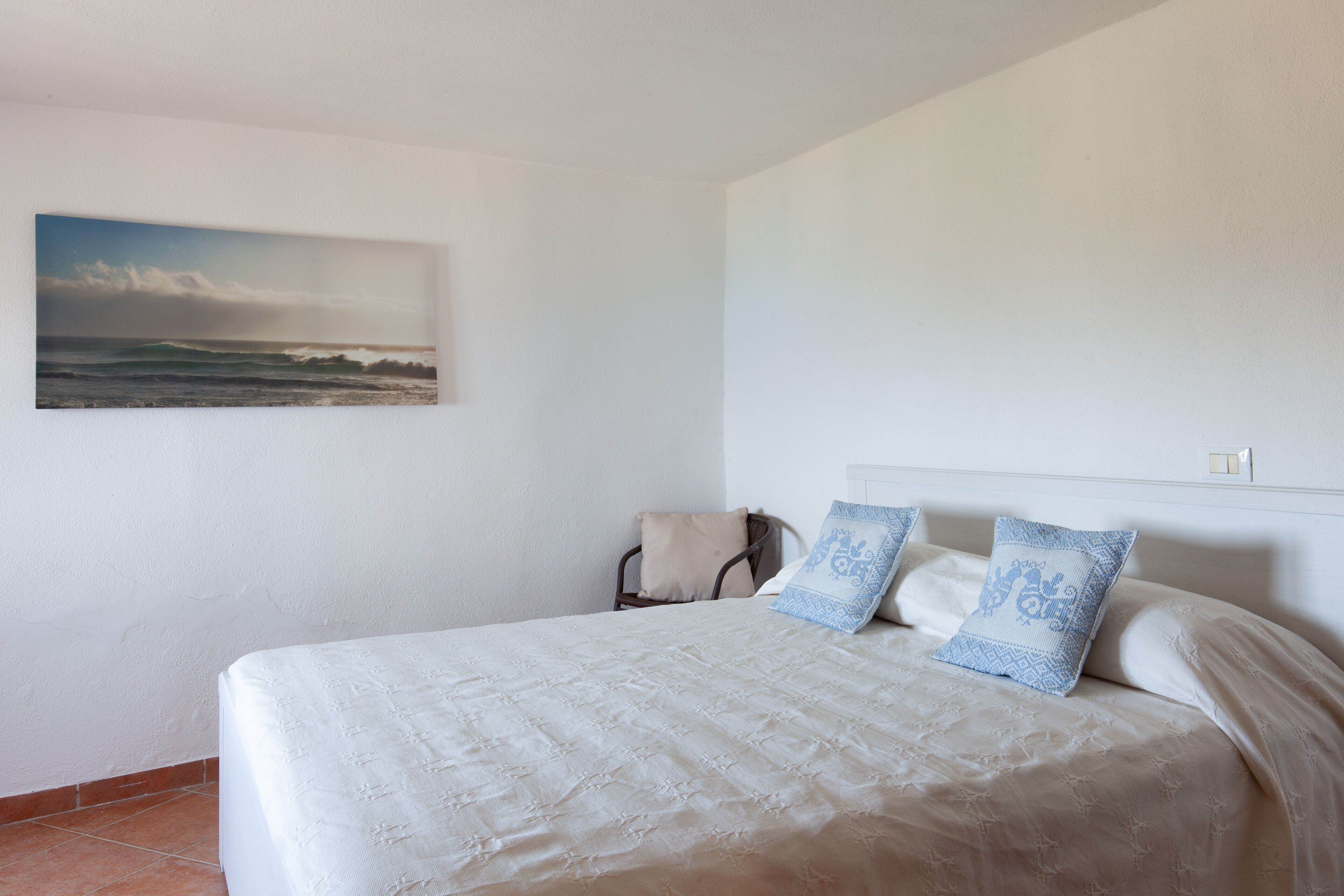 A bedroom with a bed, two pillows, a chair, and a painting of a beach scene on the wall.