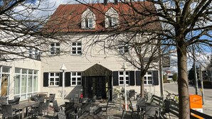 A building with a red-tiled roof, a covered entrance, and outdoor seating with chairs and tables. - Upper-House (Aystetten)