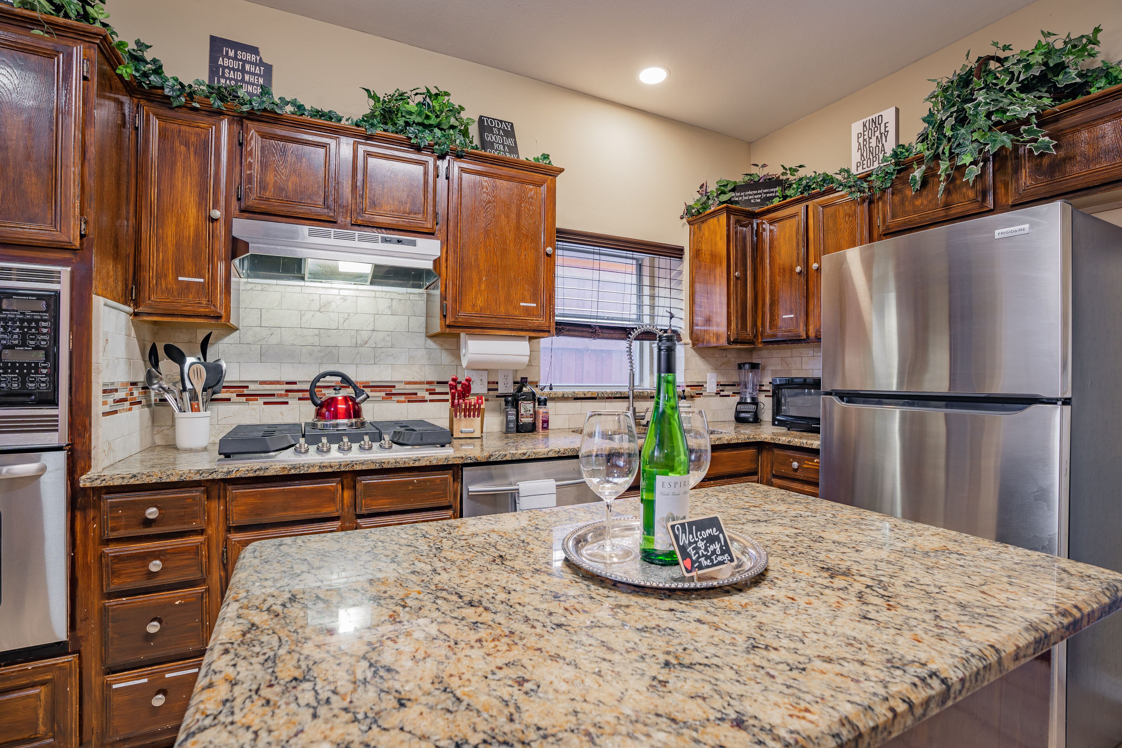 A kitchen with wooden cabinets, a stainless steel refrigerator, granite countertops, and a welcome sign.