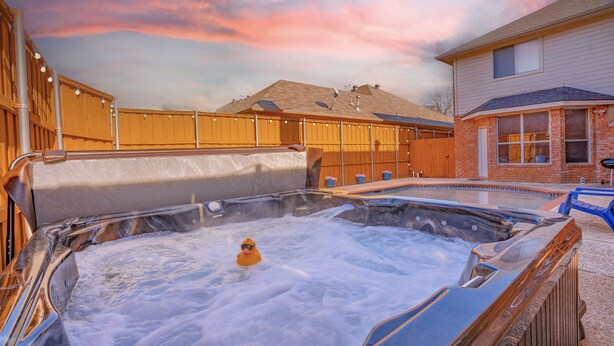 Un jacuzzi con una persona adentro, rodeado por una valla de madera y una casa al fondo.
