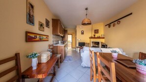 A spacious living area with a wooden dining table, a sofa, and a TV. There is a kitchen in the background with wooden cabinets and a hanging light fixture. - La Casetta dei Gerani (Montemarano)