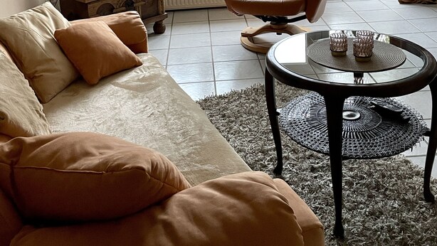 A living room with a sofa, armchair, and coffee table. There is a TV mounted on the wall, a chandelier, and a window with curtains.