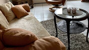 Ein Wohnzimmer mit Sofa, Sessel und Couchtisch. Ein Fernseher ist an der Wand montiert, eine Chandelüster hängt von der Decke und ein Fenster ist mit Vorhängen verziert.