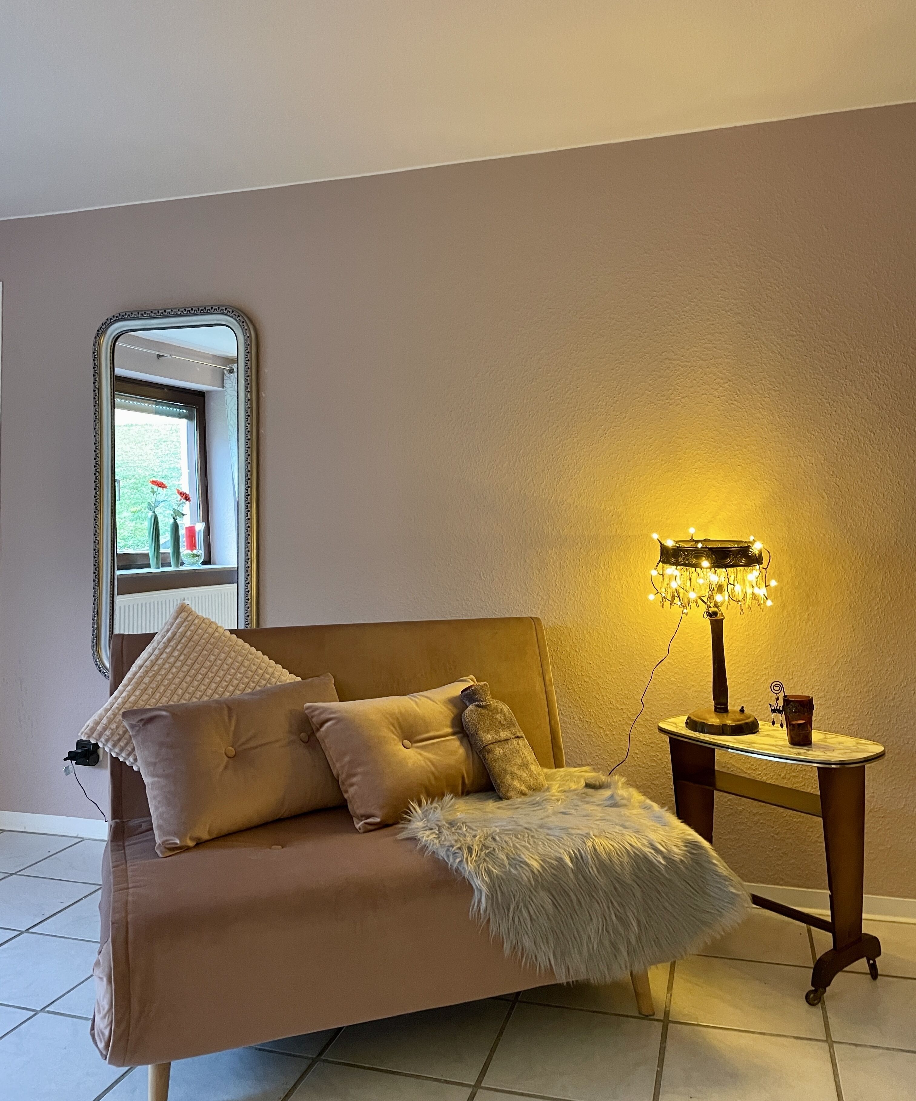 Ein gemütliches Zimmer mit einer Couch, einem kleinen Tisch mit einer Lampe und einem Fenster mit Blumen.