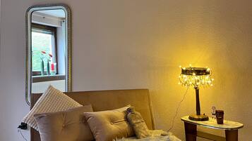 Ein gemütliches Zimmer mit einer Couch, einem kleinen Tisch mit einer Lampe und einem Fenster mit Blumen.