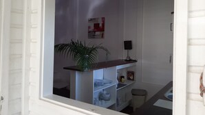 Une pièce avec un mur en briques blanches, une table en bois, une plante en pot, une étagère avec de la vaisselle et une petite poubelle.