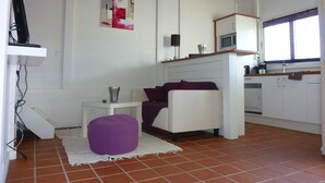 Un espace de vie compact comprenant un coin cuisine, un canapé blanc, un pouf violet et un sol carrelé.