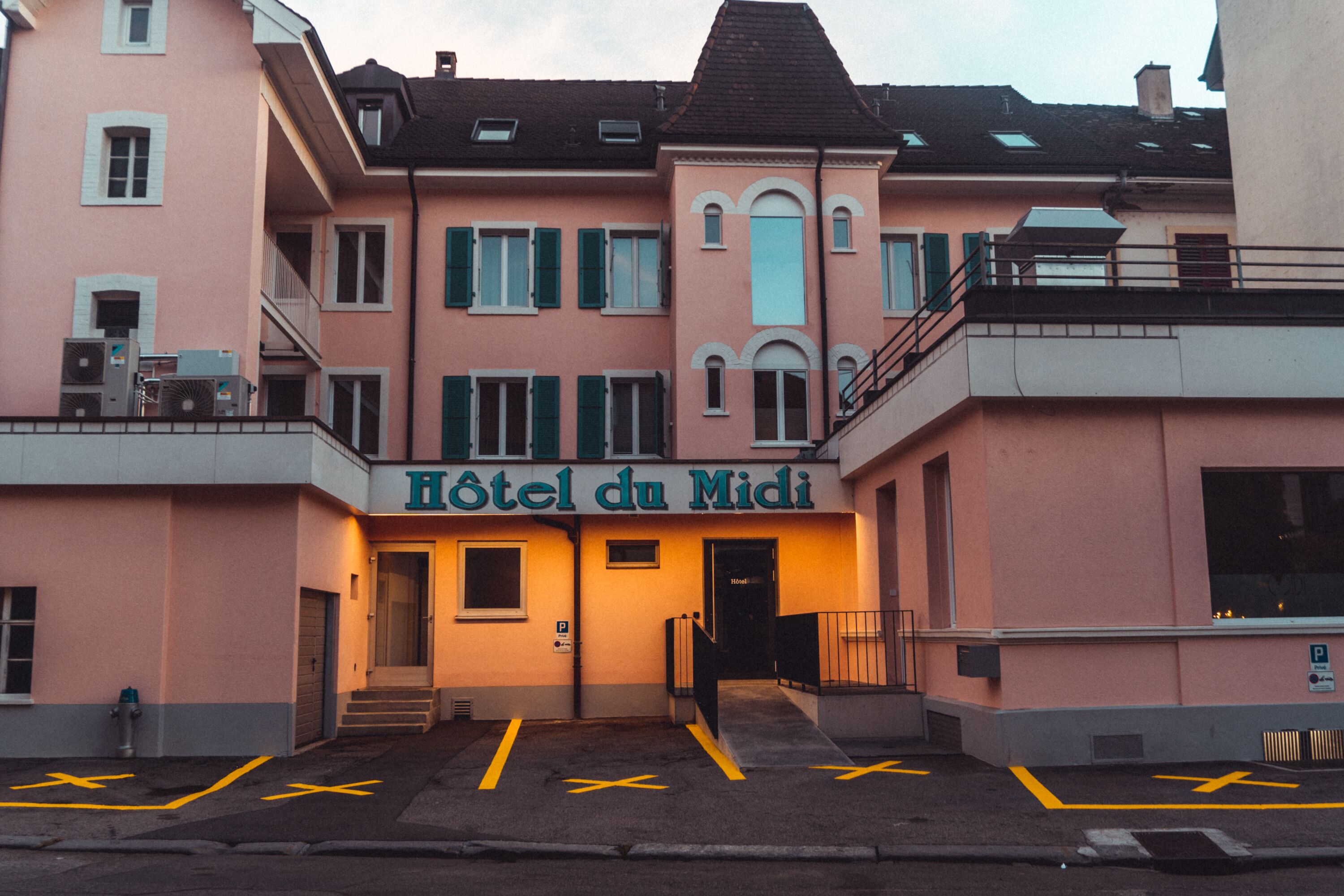 Ein Hotel namens "Hotel du Midi" mit einer rosafarbenen Fassade, zahlreichen Fenstern und einem Parkplatz mit eingezeichneten Stellplätzen.