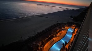 Blick auf einen Strand mit einer langen Seebrücke, einem Pool mit Rutsche und einem Sonnenuntergang im Hintergrund.