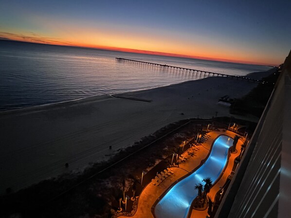Une vue sur la plage avec une longue jetée qui s’avance dans l’eau, une piscine avec un toboggan et un coucher de soleil en arrière-plan.