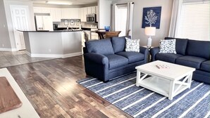 Ein modernes Wohnzimmer mit einer dunkelblauen Sofagarnitur, einem weißen Couchtisch und einem Streifen Teppich. Im Hintergrund sind eine Küche mit weißen Schränken sowie ein Essbereich mit Tisch und Stühlen zu sehen.