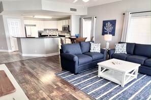 Un salon moderne avec un canapé bleu foncé, une table basse blanche et un tapis à rayures. On aperçoit à l’arrière-plan une cuisine avec des meubles blancs et un coin repas comprenant une table et des chaises.