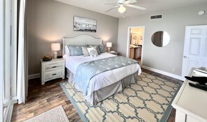 Une chambre à coucher avec un lit, des tables de chevet, un ventilateur de plafond, un tableau, un miroir et une porte.