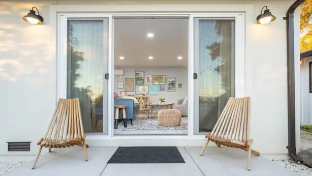 A modern house with a sliding glass door entrance, two wooden chairs outside, and a cozy interior with a sofa, table, and framed pictures on the wall.
