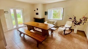 A spacious room with a wooden dining table, benches, and a white chair. There is a fireplace, a window with a view of greenery, and a door leading outside. - Green Paradise (Introdacqua)