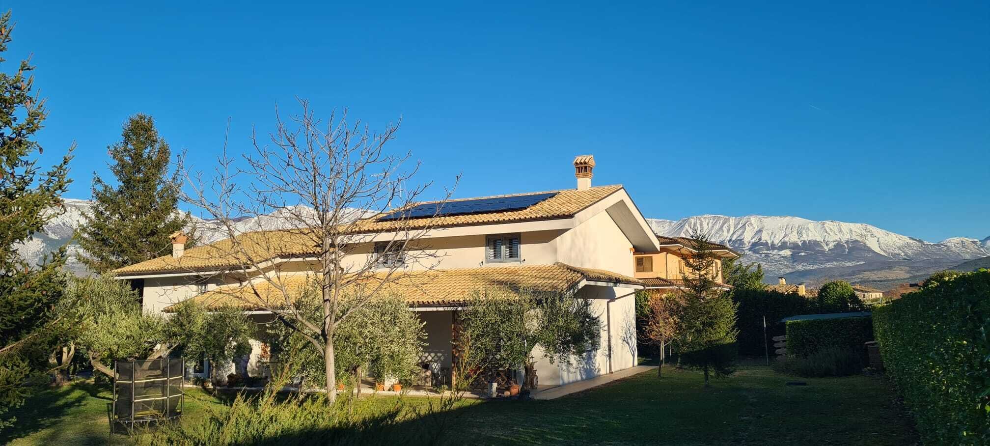 Ein zweistöckiges Haus mit Ziegeldach, umgeben von viel Grün und Bergen im Hintergrund.