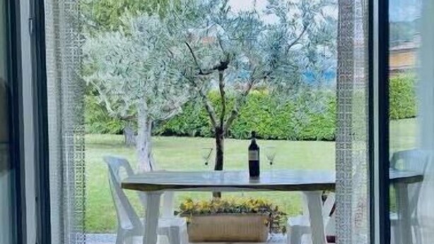 A view through a glass door into a patio area with a table and chairs, a bottle, and a garden with trees and grass. - Green Paradise (Introdacqua)