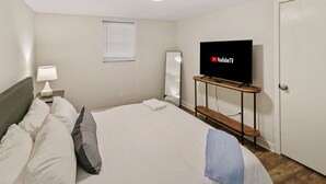 Hotellihuone, jossa on sänky, puinen jalusta, jonka päällä on televisio, yöpöytä lampun kanssa ja ikkuna, jossa on sälekaihtimet.