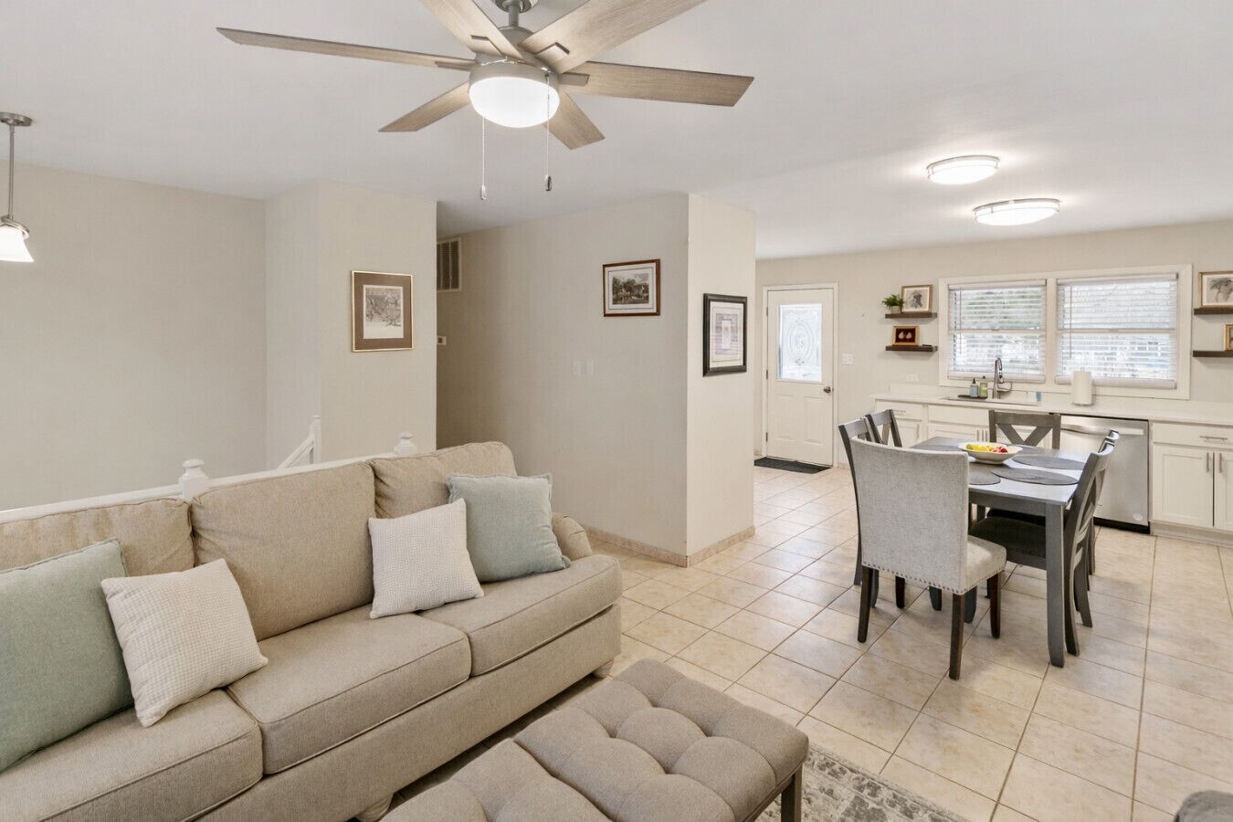 A modern living room with a beige sofa, a dining table with chairs, a ceiling fan, and a kitchen area with a window.