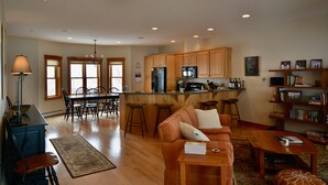 A spacious living room with a sofa, coffee table, and a rug. There is a kitchen area with wooden cabinets and a refrigerator.