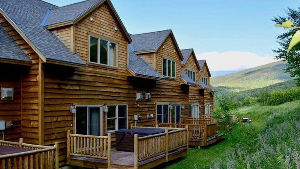 A row of log cabin-style cabins with wooden decks and large windows, set in a lush, green landscape.