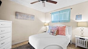 Ein Schlafzimmer mit einem Bett, einem Deckenventilator, einer Kommode, einem Fenster mit blauer Gardine und einem Wandbild in einem Rahmen.