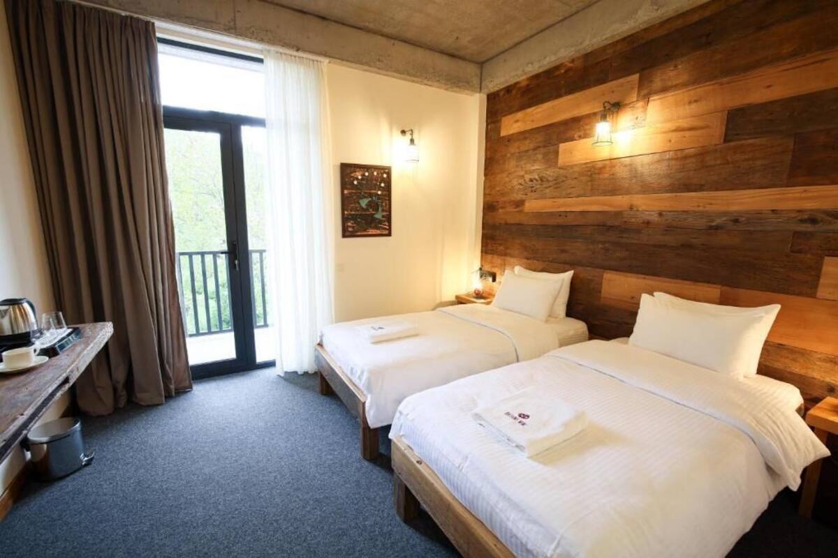 A hotel room with two beds, a wooden headboard, a desk with a kettle, and a view through a glass door.