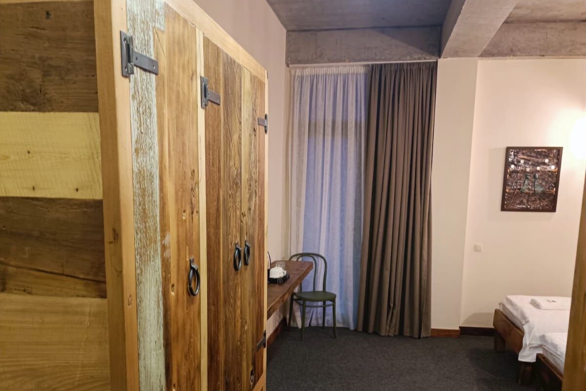 A room with a large wooden wardrobe, a small table with a chair, a bed, and a curtain.