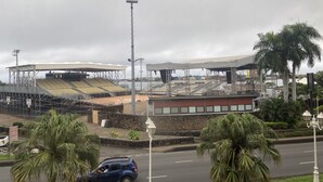 Een stadion met meerdere zitplaatsen en een podium, omringd door palmbomen en een weg met een passerende auto.