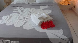 Een net opgemaakt bed met een bloemenbedekker, witte kussens en een opgerolde handdoek met rode bloemen.