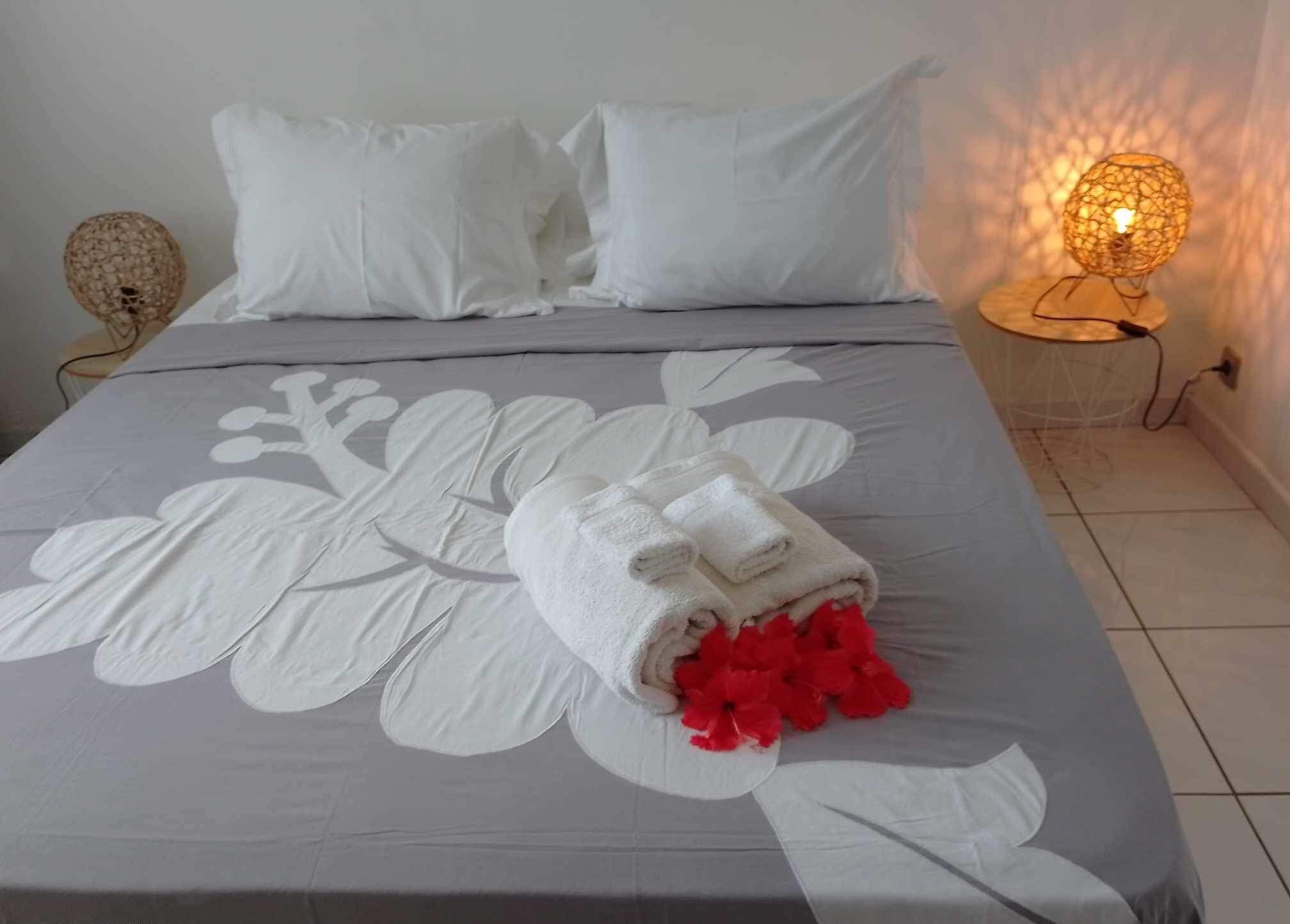 A neatly made bed with a floral bedspread, white pillows, and a rolled towel with red flowers.