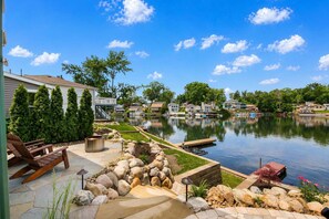 A lakeside patio with a wooden deck, a stone fire pit, and a view of a residential area across the water. - Lake Front - Kayaks - Huge Game Room - Sleeps 8 (Akron)